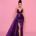 A woman stands confidently against a pink background, wearing the NP162, a sleek, deep purple satin dress with a thigh-high slit from the Best Sunshine Coast Dress Shop. Her hair is styled in an elegant updo, and she is wearing matching earrings and strappy high heels. Angels Formal Wear