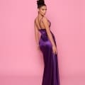 A woman with an elegant updo wears the NP162 floor-length, fitted purple satin gown with thin straps, posing against a solid pink background. She looks over her shoulder with a confident expression, showcasing the sleek design from Best Sunshine Coast Dress Shop and her radiant look. Angels Formal Wear