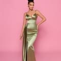 Styled by the Best Sunshine Coast Dress Shop, a woman poses against a pink background in NP162, a form-fitting metallic green gown with a deep V-neckline and a dramatic drape on one side. Her hair is elegantly styled in an updo, complemented by statement earrings. Angels Formal Wear