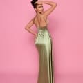 A woman stands with her back turned, showcasing the NP162 gown from the Best Sunshine Coast Dress Shop. The long, silky dress features a light green hue, a deep open back, and thin straps. Her hair is styled in an elegant updo against a solid pink background. Angels Formal Wear