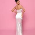 A woman with an elegant updo poses against a pink background, showcasing the back of her strapless RITA NP165 white satin gown. The dress features a fitted bodice and a full-length skirt that flows gracefully to the floor. Her hand rests on her hip as she looks over her shoulder, embodying the elegance found at the Best Sunshine Coast Dress Shop. Angels Formal Wear