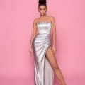 A woman poses confidently against a pink backdrop, wearing the RITA NP165 from Best Sunshine Coast Dress Shop—a strapless, form-fitting, metallic silver gown with a high slit revealing one leg. She accessorizes with hoop earrings and nude high heels, has her hair styled in an elegant updo, and stands with her hands at her sides. Angels Formal Wear