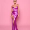 A woman wearing the ALLURE NP166, a shiny, form-fitting, pinkish-purple gown with a deep neckline and a high slit on one side, poses against a solid pink background. Styled in an updo and accessorized with statement earrings, she looks ready for any event at the Sunshine Coast Cocktail Dress Shop. Angels Formal Wear