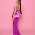 A woman with long, braided hair stands against a pink background. She is wearing the ALLURE NP166, a shiny, form-fitting magenta evening gown with spaghetti straps from the Cocktail Dress Shop on the Sunshine Coast. She glances over her shoulder towards the camera, one arm relaxed by her side. Angels Formal Wear