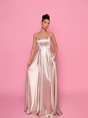 Against a pink background, a woman poses in the CHANTELLE NP168 gown from the Cocktail Dress Shop. The strapless metallic dress features a fitted bodice and flowing skirt. Her hair is styled up, complemented by earrings, with one hand elegantly holding part of her skirt and the other resting by her side. Angels Formal Wear