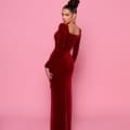 A woman stands against a pink background, posing to show her back while looking over her shoulder. She wears the form-fitting, long-sleeved deep red gown, NP169, from Cocktail Dress Shop and has an intricate updo hairstyle. She accessorizes with large, round earrings. Angels Formal Wear