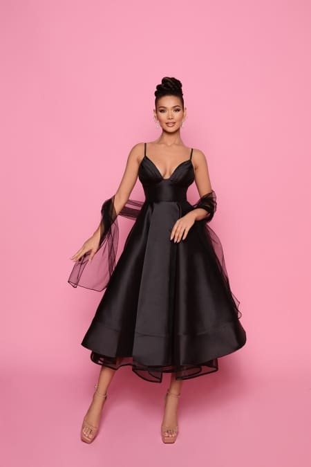 A woman stands against a pink backdrop wearing the NP175 sleeveless black cocktail dress with spaghetti straps and a full skirt. She has a sheer black shawl draped over her arms and is wearing high heels. Her hair is styled in an elegant updo, accessorized with earrings, perfect for Race Day on the Sunshine Coast. Angels Formal Wear