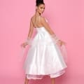 A woman poses against a pink background, wearing the NP175 - a white sleeveless A-line midi dress with a double-strap design. She accessorizes with statement earrings, high-heeled shoes, and a sheer shawl draped elegantly over her arms. Perfect for Race Day Dresses on the Sunshine Coast, she has her hair styled in an updo. Angels Formal Wear