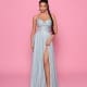 A woman stands against a pink background wearing the LEXI NP176, a shimmery light blue strapless gown with a sweetheart neckline and high slit, ideal for Race Day Dresses. She has long braided hair, statement earrings, and neutral-colored high heels. Angels Formal Wear