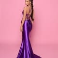 A woman stands against a pink background, wearing the RYDER NP180, a floor-length purple satin gown featuring an open back with crisscross lace-up detailing. Sporting a braided ponytail, she poses slightly to the side, glancing over her shoulder—capturing the elegance reminiscent of Sunshine Coast sunsets. Angels Formal Wear