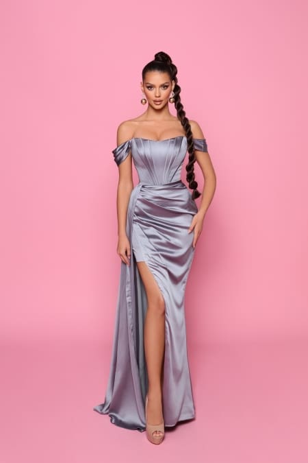 Against a pink background, a woman elegantly dons the NP185—a striking, off-the-shoulder silver gown featuring a thigh-high slit that makes it perfect for Sunshine Coast events. Her long braided hair cascades down as she accessorizes with large earrings and high-heeled sandals, creating an ideal ensemble for Race Day. Angels Formal Wear