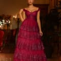 A woman stands in an elegant, dimly lit room wearing the TE206, a long, off-the-shoulder, tiered dress in deep wine color with sparkling details. Her hair is styled neatly as she holds one side of the dress. Chandeliers and mirrors adorn the background, showcasing formal wear online from Australia. Angels Formal Wear