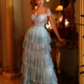 A woman stands beside an ornate wooden table and a large mirror, wearing the off-shoulder, tiered TE206 gown adorned with sequins. The setting includes tall, elegant columns and warm lighting, creating a luxurious ambiance—perfect for those searching for formal wear online in Australia. Angels Formal Wear