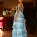 A woman stands in an elegant room, wearing the TE206 blue off-the-shoulder gown adorned with tiered ruffles and sparkling details. She looks over her shoulder toward the camera. The room is dimly lit with ornate decorations and warm lighting, creating a luxurious ambiance reminiscent of Formal Wear Online Australia selections. Angels Formal Wear