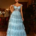 A woman stands indoors in an elegant setting, wearing the TE206, a floor-length tiered light blue gown with glittery details sourced from Formal Wear Online Australia. The bodice is form-fitting with off-the-shoulder straps. The room features ornate mirrors, plants, and warm lighting from chandeliers. Angels Formal Wear