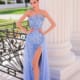 A woman in a strapless blue gown with intricate beadwork stands beside a decorative wrought-iron railing and large white columns. The TK118, from a Dress Shop on the Sunshine Coast, has a high slit revealing her left leg. She is standing outdoors in a sunny setting. Angels Formal Wear