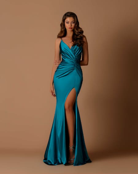 A woman stands against a neutral brown background, wearing a teal satin evening gown from AQUARIUS NBM1036. The dress features a V-neck, gathered bodice, and a high slit on the right leg. She has long, wavy hair and is posing with one hand on her hip and the other resting by her side. Angels Formal Wear