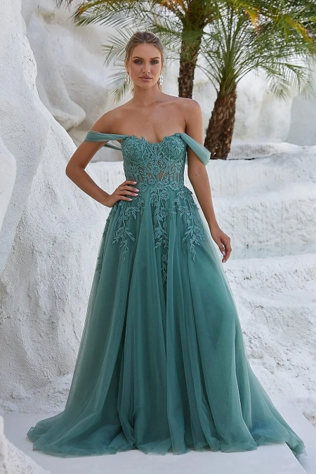 A woman stands in front of white, rocky terrain with palm trees, showcasing evening wear. She wears a green, off-the-shoulder gown with a lace bodice and flowing skirt from Dress Shop Sunshine Coast. Her hair is styled up, and she has a calm expression. The dress contrasts beautifully with the natural background. Angels Formal Wear