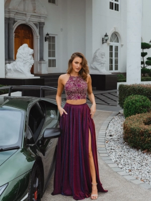 A woman stands confidently beside a sleek green sports car outside a grand white building in Manchester. She is wearing a maroon two-piece outfit: a glittery crop top and a matching long skirt with a high slit, accessorized with beige high-heeled sandals and holding her MANCHESTER PO893 handbag. Angels Formal Wear