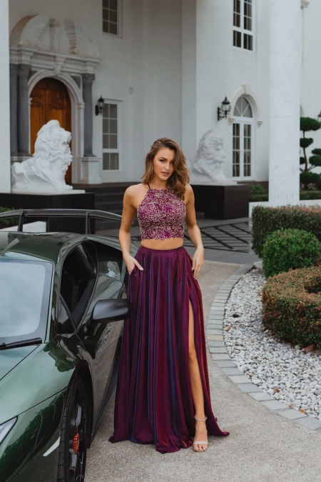 A woman stands confidently beside a sleek green sports car outside a grand white building in Manchester. She is wearing a maroon two-piece outfit: a glittery crop top and a matching long skirt with a high slit, accessorized with beige high-heeled sandals and holding her MANCHESTER PO893 handbag. Angels Formal Wear