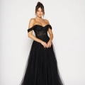 A woman stands against a plain white background wearing an elegant off-the-shoulder black gown with intricate lace detailing and a sheer, flowing skirt. Her hair is styled in an updo with loose strands framing her face, and she holds her hands together in front. Buy INNES PO2462 online for the perfect look. Angels Formal Wear