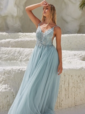 A woman wearing a flowing, light blue, lace-trimmed gown with spaghetti straps stands against a textured white stone background. She is posing with one hand shielding her face from the sun, her gaze directed upwards. The scene appears elegant and serene, just like those you find when you buy COVE PO2453 online. Angels Formal Wear