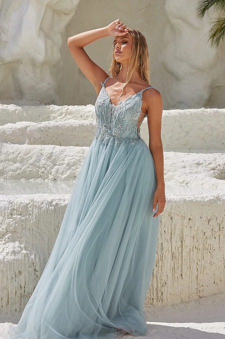 A woman wearing a flowing, light blue, lace-trimmed gown with spaghetti straps stands against a textured white stone background. She is posing with one hand shielding her face from the sun, her gaze directed upwards. The scene appears elegant and serene, just like those you find when you buy COVE PO2453 online. Angels Formal Wear