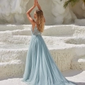 A woman with long blonde hair stands with her back to the camera, wearing a flowing, light blue gown with intricate detailing on the bodice. She is raising her arms gracefully above her head, surrounded by white, rocky terrain and green foliage. Discover similar styles when you buy COVE PO2453 online. Angels Formal Wear