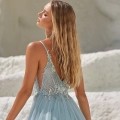 A woman with long blonde hair is wearing a flowing, light blue dress with delicate lace detailing on the back. She stands outdoors on a white, textured surface, with her head tilted back and eyes closed, basking in the sunlight. Explore similar styles and buy COVE PO2453 online to enhance your wardrobe. Angels Formal Wear