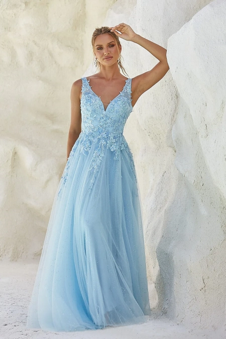 A woman stands against a textured white wall, wearing a flowing light blue gown adorned with floral appliqués and a deep V-neckline. The dress features a sheer overlay with delicate sparkles, and the woman has a relaxed, elegant pose with one hand resting on the wall. Buy MIRA PO2440 online for similar styles. Angels Formal Wear