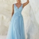 A woman stands against a textured white wall, wearing a flowing light blue gown adorned with floral appliqués and a deep V-neckline. The dress features a sheer overlay with delicate sparkles, and the woman has a relaxed, elegant pose with one hand resting on the wall. Buy MIRA PO2440 online for similar styles. Angels Formal Wear