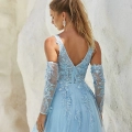 A woman stands with her back to the camera, showcasing a flowing light blue MIRA PO2440 adorned with floral lace details and sheer long sleeves. She has blonde hair tied in a high ponytail and is wearing large statement earrings. The background features pale rock formations, making it an ideal scene to inspire you to buy formal dresses online. Angels Formal Wear