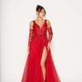 A person is wearing an elegant red evening gown with a deep V-neckline and intricate floral embellishments. The MIRA PO2440, available when you buy formal dresses online, features sheer long sleeves and a high slit on one side, revealing a leg. The person has their hair styled in an updo and is smiling. Angels Formal Wear