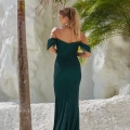 A woman with blonde hair in a ponytail is wearing an off-shoulder, fitted, dark green gown. She stands with her back to the camera against a backdrop of white rock and lush green palm trees. The gown's train elegantly drapes over the ground. Buy CARLOW PO2465 online for a stunning look like this. Angels Formal Wear