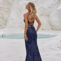 A woman in a blue, backless, lace-up gown from AMA PO2454 stands on a white sandy area with rocky formations in the background. Her long, wavy hair flows down her back as she faces away from the camera, gazing at a small pool of water. Angels Formal Wear