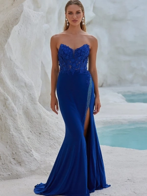 A woman stands gracefully in front of a rocky, white-walled background with turquoise water. Wearing a strapless, royal blue gown with intricate lace detailing and a thigh-high slit from BO PO2452, she accessorizes elegantly with long earrings. Angels Formal Wear