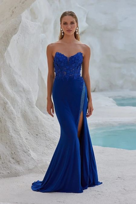 A woman stands gracefully in front of a rocky, white-walled background with turquoise water. Wearing a strapless, royal blue gown with intricate lace detailing and a thigh-high slit from BO PO2452, she accessorizes elegantly with long earrings. Angels Formal Wear