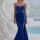 A woman stands gracefully in front of a rocky, white-walled background with turquoise water. Wearing a strapless, royal blue gown with intricate lace detailing and a thigh-high slit from BO PO2452, she accessorizes elegantly with long earrings. Angels Formal Wear