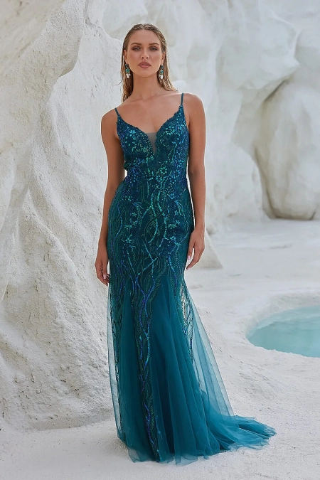 A woman stands against a textured, white rocky background, wearing a teal, sleeveless, floor-length gown adorned with intricate beadwork. Her long hair is styled loosely, and she wears matching teal earrings from OCEANE PO2461. A small pool of water is visible in the background. Angels Formal Wear