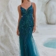 A woman stands against a textured, white rocky background, wearing a teal, sleeveless, floor-length gown adorned with intricate beadwork. Her long hair is styled loosely, and she wears matching teal earrings from OCEANE PO2461. A small pool of water is visible in the background. Angels Formal Wear