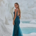 A woman in a sleeveless, backless teal gown with intricate beading from the OCEANE PO2461 stands on a snowy white landscape, looking over her shoulder. The background features icy formations and a blue pool of water, creating a striking contrast with her elegant dress. Angels Formal Wear