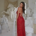 A woman in a red, backless, sequin gown RAINEY PO2451 poses gracefully with her hand on a white sculpture in an art studio. The studio is filled with various white sculptures, and a wooden ladder is visible on the left side. Angels Formal Wear