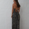 A woman smiles while facing away from the camera, wearing a black, floor-length, sequined dress with an intricate strappy back design. The background is a plain, light-colored wall, showcasing elegant options available through RAINEY PO2451. Angels Formal Wear