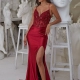 A woman in a red, floor-length TARNI PO2445 from Online Formal Wear Australia with a high slit is standing beside a white sculpture. The dress has a lace bodice with thin straps and a satin skirt. She is smiling, resting one hand on the sculpture. The background features several other sculpture pieces and white decor. Angels Formal Wear