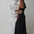 A woman in a strapless black evening gown with cutouts on the side, sourced from Online Formal Wear Australia, poses with her back to the camera next to a white marble statue of a man. She looks over her shoulder towards the camera, showcasing her long dark hair and elegant dangling earrings. The gown is identified as MAR PO2439. Angels Formal Wear