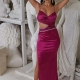 A woman poses in a form-fitting, sleeveless magenta SARITA PO2443 gown with cut-out details at the waist. She stands in front of a background filled with classical-style white sculptures and statues. She has her hands raised, resting behind her head, and wears drop earrings and high heels—a perfect look from Online Formal Wear Australia. Angels Formal Wear