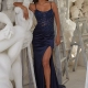 A woman in a navy blue gown with a thigh-high slit and intricate beadwork on the bodice, tagged as GAL PO2467, leans against a gray ladder in an art studio filled with white sculptures. She is wearing white platform sandals and has her hand resting on one of the sculptures. Angels Formal Wear