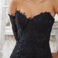 A woman wearing the elegant TALISE PO2468 off-the-shoulder black lace dress with intricate floral details and sequins. The dress, featuring a sweetheart neckline, and she has a pearl earring. Her hair cascades over one shoulder against a softly blurred background. Angels Formal Wear