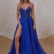 A woman stands on sand against a textured beige wall, wearing the AQUA PO2447 with a deep V-neckline and intricate beadwork. The dress has a thigh-high slit, and she has long blonde hair styled in loose waves. She is looking straight ahead with a neutral expression. Angels Formal Wear