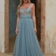 A woman stands in front of a rustic doorway wearing a floor-length, sleeveless gown reminiscent of the TALLULAH PO2469. The top of the dress is embellished with intricate beadwork, and the flowing skirt is made of soft, airy fabric in a muted blue-green color. She has long, wavy hair. Angels Formal Wear